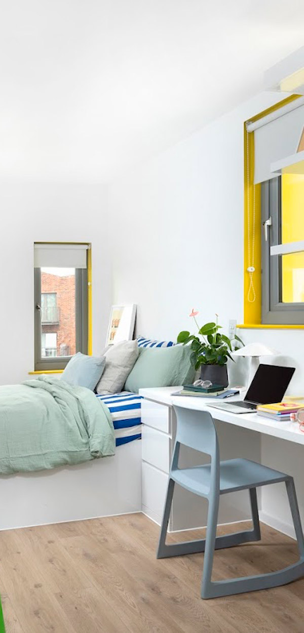 A stylish bedroom featuring a green bedspread, striped pillows, a desk with a laptop, and a window with yellow accents, creating a cozy and vibrant atmosphere. Perfect for modern living.