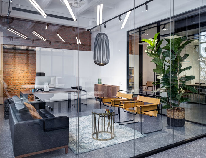 Modern office interior featuring a sleek black sofa, wooden chairs, a glass partition, and a decorative plant, designed for a stylish workspace.