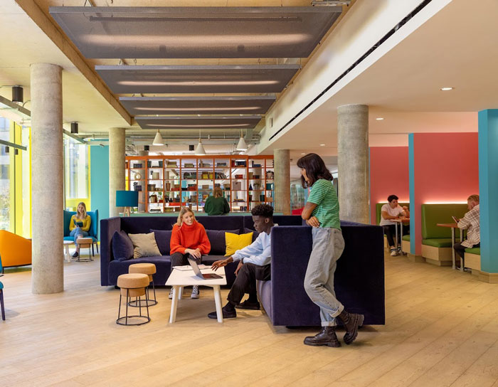 A modern, vibrant lounge area with people engaging in conversation and collaborative work around a central table, featuring stylish furniture and colorful walls. Ideal for socializing or studying.