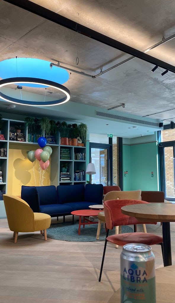 Contemporary lounge area with colorful furniture, balloons, bookshelves, and a drink can. Cozy seating and bright lighting create an inviting atmosphere.