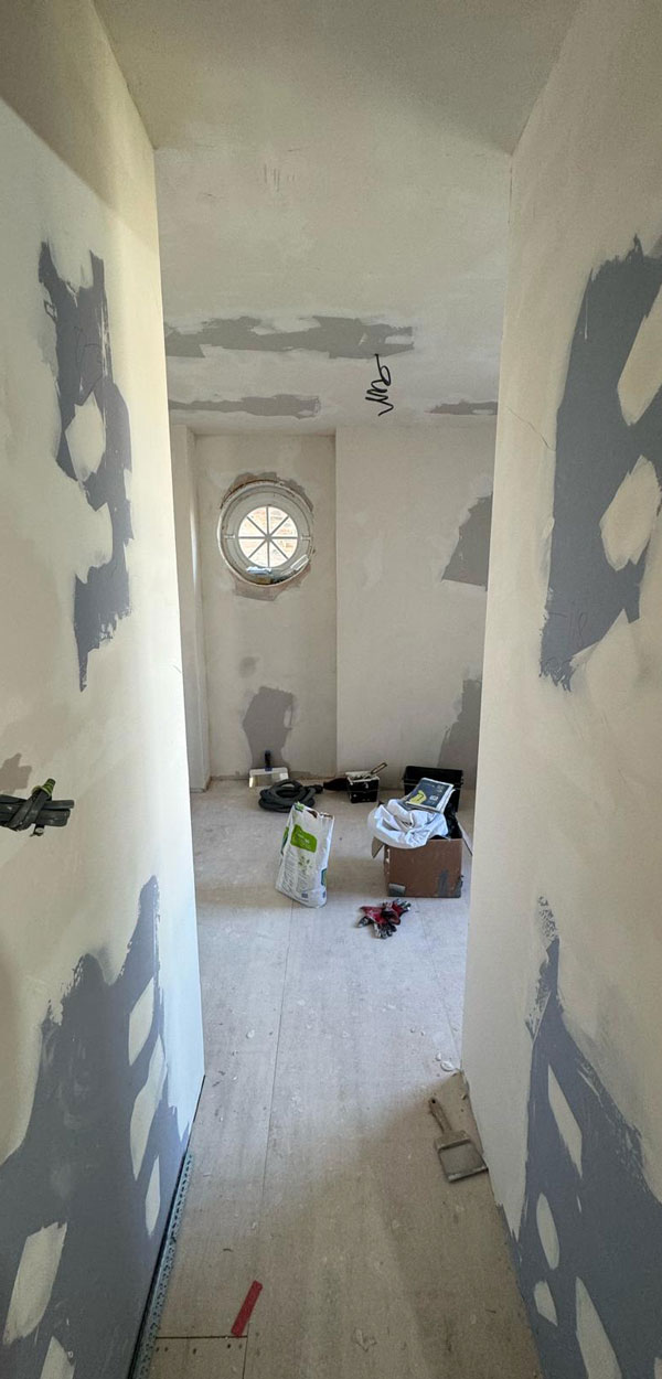 Interior view of a partially finished hallway with unpainted walls featuring gray patches and a round window at the end, along with scattered construction materials on the floor.