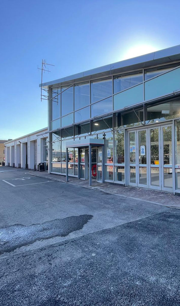 Modern commercial building entrance with large glass windows, showcasing a clear blue sky and a nearby parking area.