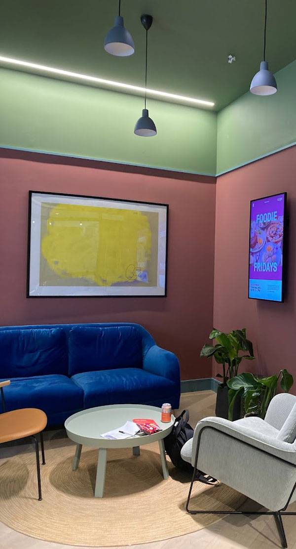 Cozy lounge area featuring a blue sofa, a round table with magazines, an armchair, decorative plants, and an art piece on the wall. The space has dim lighting with green and pink walls, creating a comfortable and inviting atmosphere for relaxation or socializing.