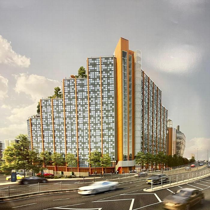 A modern multi-story building with large glass windows and orange accents, surrounded by greenery and traffic on a busy road.