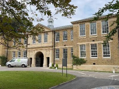 A historic brick building with large windows and a clock tower, surrounded by greenery and a van parked in front, showcasing classic architectural style.