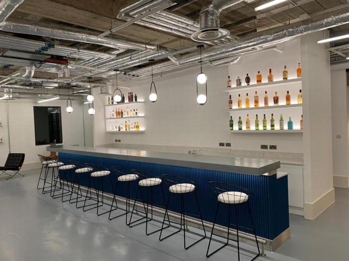 Modern bar interior featuring a blue counter with stools, illuminated shelves displaying various bottles, and industrial ceiling design.